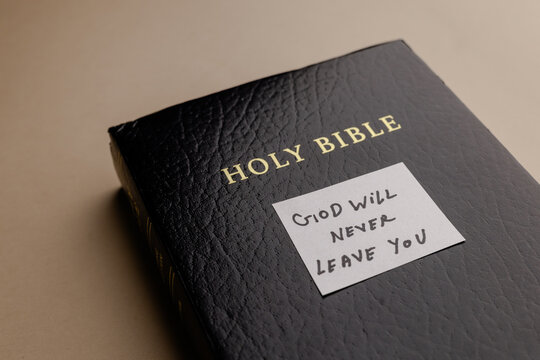 A black leather holy bible book lie on a light surface with a white note paper stuck on cover with a handwritten quote about faith and hope