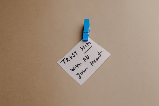 A small white note with handwritten Christian quote attached by a blue clothespin to a neutral background, inspiring trust and faith message