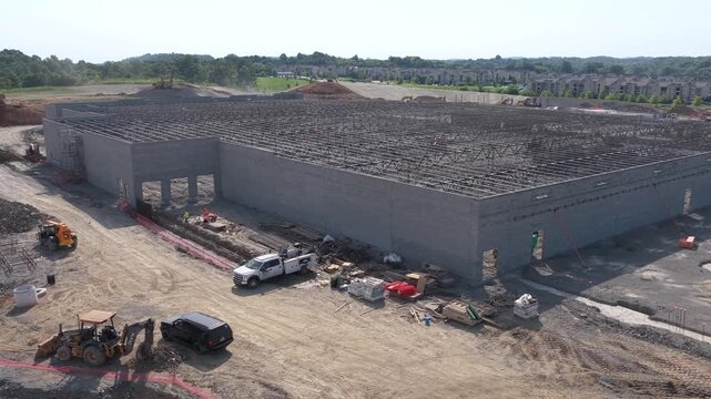 A tilt up concrete logistics warehouse stands half built in Mount Juliet, Tennessee, open steel roof joists over bare pads with a backhoe and pickups parked on the dirt. Aerial approaching shot