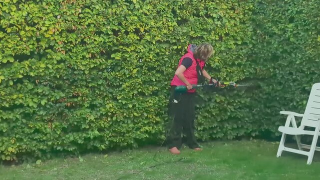 Young woman trims tall bushes with an electric trimmer in a garden setting