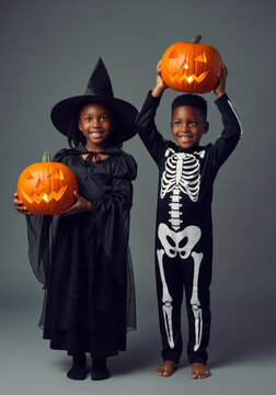 Girl dressed as a witch and boy dressed as a skeleton hold carved pumpkins. They pose in costume against a gray studio background. Halloween family celebration concept.