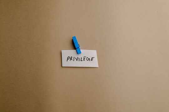 A small white note with the word privilege written on it held by a blue wooden clothespin on a plain beige background conceptual shot