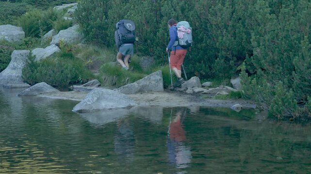 Family with Child Hiking Along Shaded Mountain River at Sunrise. Low angle motorized camera tracking past multi-generational family walking away. Calm alpine river with sunrise reflections and ...