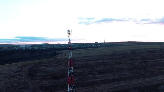 Red-and-white cellular communications tower with panel antennas in a drone orbit near a river. Telecommunications infrastructure, mobile broadband and regional connectivity