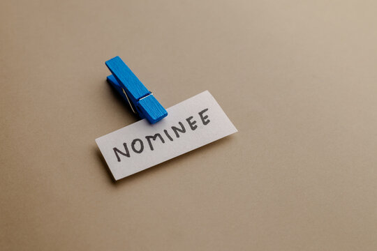 A small white paper slip with the word nominee written in black ink, attached to a blue wooden clothespin on a neutral beige background