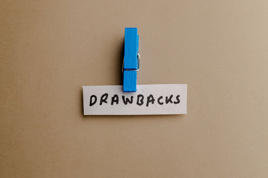 A small paper strip with the handwritten text drawbacks held by a bright blue wooden clothespin on a plain beige surface background style