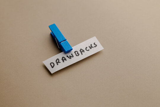 A close up view of a paper slip with the word drawbacks written on it held by a blue wooden peg clip isolated on a beige background