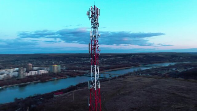 Red-and-white cellular communications tower with panel antennas in a close drone orbit. Mobile broadband and microwave backhaul network infrastructure
