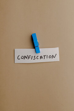 A small white paper strip with the handwritten word confiscation attached by a bright blue wooden clothespin against a solid tan background