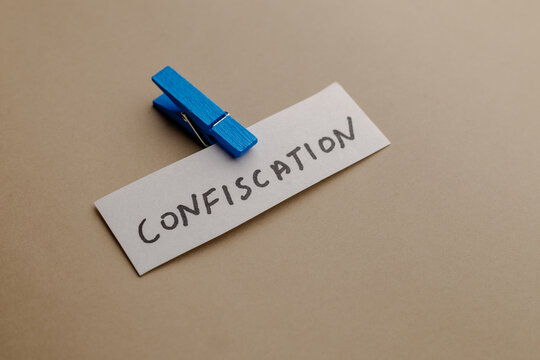 A close up view of a white paper strip with handwritten text word confiscation clipped with a blue wooden clothespin on a beige background
