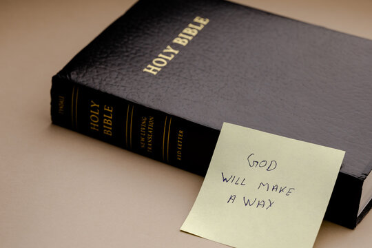 A black holy bible book rests on a soft surface next to a yellow sticky note with handwritten text about faith, hope, and divine guidance