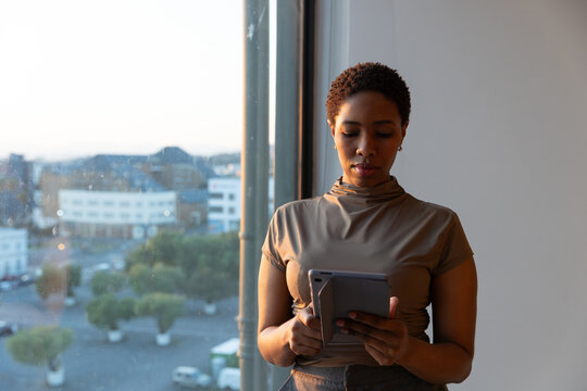 African American woman standing by large window holding tablet and working inside office