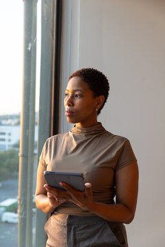 African American woman standing at office window holding tablet, wearing tan top and gray trousers