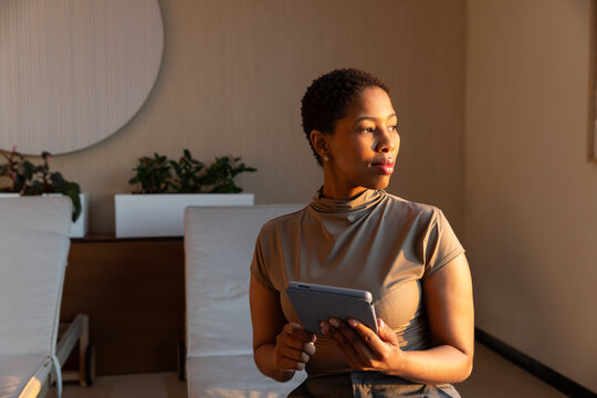 Adult woman sitting on lounge chair by console with planters holding tablet gazing right