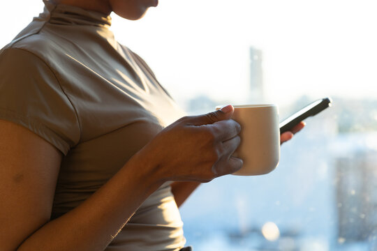 African woman holding ceramic mug and smartphone leaning by big window wearing fitted high neck top