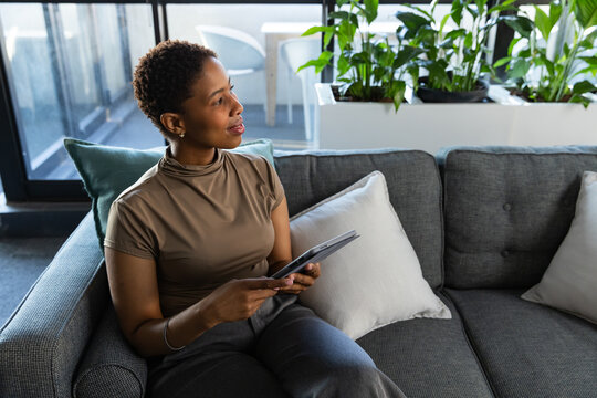 African American woman sitting on gray sectional sofa in lounge, holding tablet, looking right