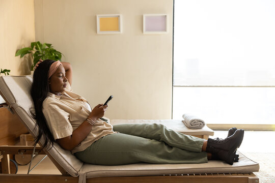 Adult African woman reclining on lounge chair at home, holding smartphone, wearing pink headband