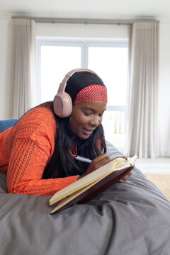 African American woman lying on bed writing with pen in journal wearing orange knit pink headphones