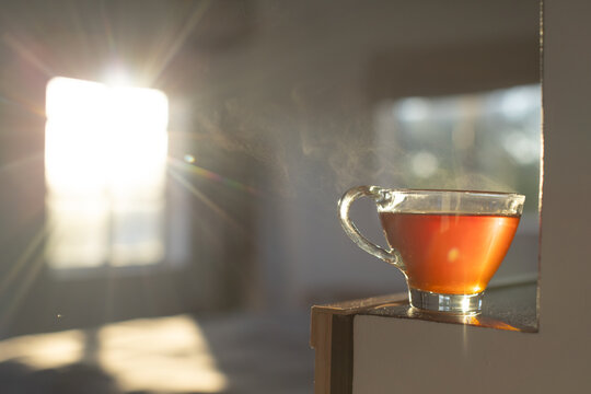 Clear glass cup is steaming on wooden ledge by large window, sunlight rays creating warm bokeh