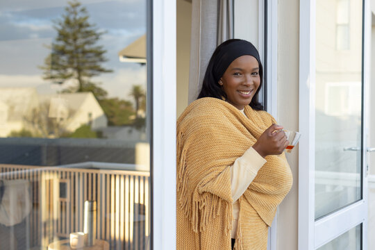 African American woman wearing mustard shawl smiling, holding glass cup on balcony, copy space