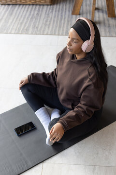 African American woman in brown sweater meditating on gray mat at home with phone and headphones