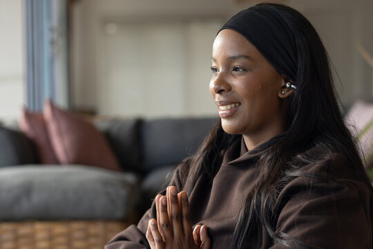 African American woman praying in living room with hands pressed brown hoodie dark headband earbud