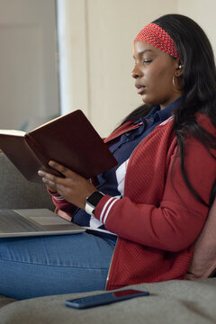 African American adult woman sitting on gray sofa in red sweater reading with laptop and smartphone
