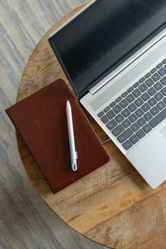 Closed brown leather notebook is resting on round wooden table near silver laptop, white pen