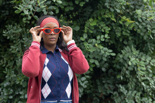 African American woman adjusting red cat-eye shades while wearing argyle top red jacket by hedge