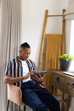 African American man wearing striped shirt sitting in armchair using tablet near window with plant