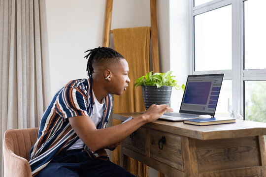 African American man leaning typing at desk wearing striped shirt with laptop, earbud, mouse, plant