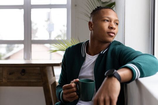 African American man sitting at window holding green mug, in green jacket and smartwatch
