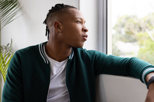 Adult African man sitting, resting arm on windowsill, gazing out window, wearing dark green jacket