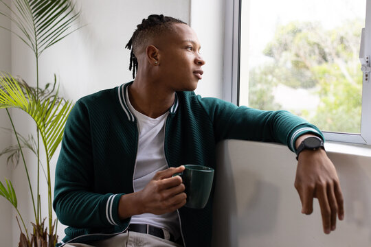 African American man in green jacket sitting on window ledge holding dark green mug