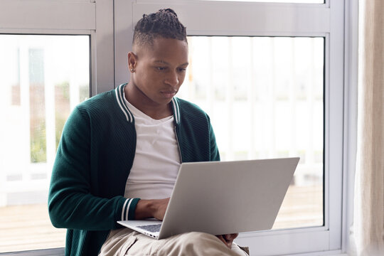 Adult African American man wearing dark green jacket by glass sliding door at home using laptop