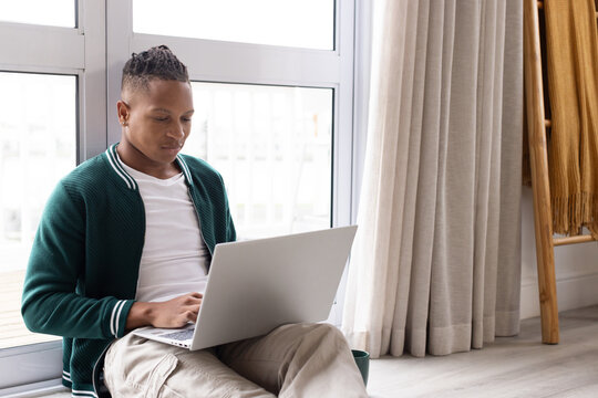 African American man in green jacket sitting at home by glass door using laptop with mug