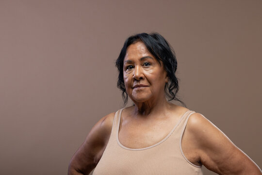 Senior female standing with hands on hips, wearing beige tank top in studio with beige backdrop
