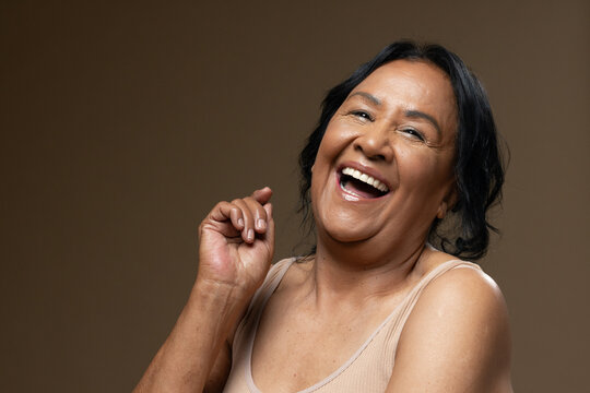 Senior African American woman laughing, hand raised in studio warm brown backdrop wearing beige top