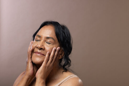Senior African American woman pressing hands to cheeks with eyes closed wearing beige top in studio