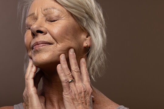 Senior woman touching neck and lower face in studio showing gold jewelry, tank strap, wrinkles