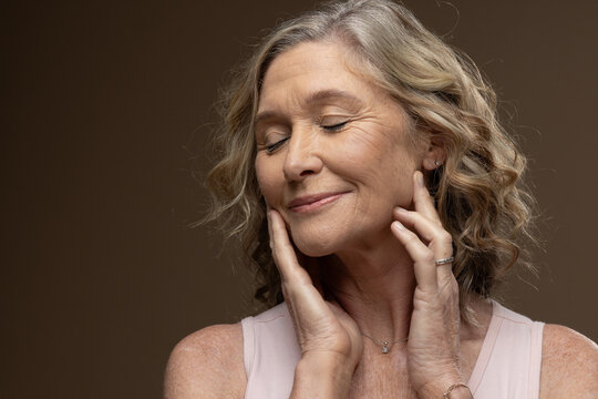 Senior woman touching jaw and neck, wearing pale sleeveless top and pendant necklace in studio