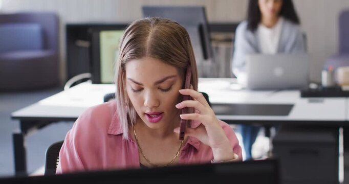 African American woman answering smartphone call for work while reading laptop at shared desk