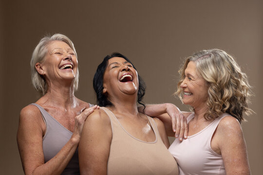 Diverse female friends laughing, leaning in studio with beige backdrop and sleeveless tops