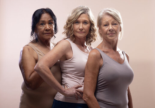 Mature female trio posing in studio with beige backdrop, soft lights, sleeveless tops