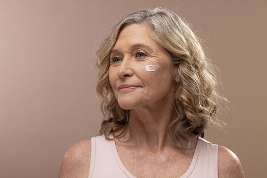 Senior woman posing in studio with pale pink tank top and skincare cream on cheek