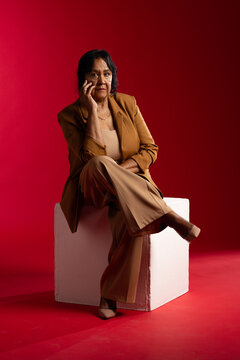 Senior African woman posing on white cube pedestal in studio with red backdrop and tan suit