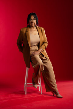 Senior African American woman sitting on white wooden stool wearing tan blazer and heels in studio