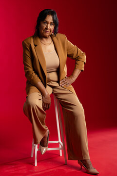 Senior African American woman sitting on white wooden stool posing in studio wearing tan blazer