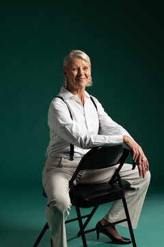 Senior woman sitting on backward black folding chair in studio, wearing suspenders and gold hoops