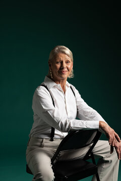 Senior woman sitting on backward black folding chair in studio wearing white shirt and suspenders